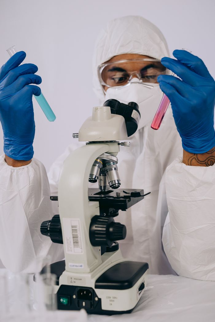 Scientist in protective suit examining test tubes in a lab setting with a microscope.