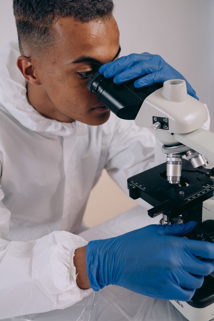 Scientist wearing PPE examines sample under a microscope indoors, focused on research.